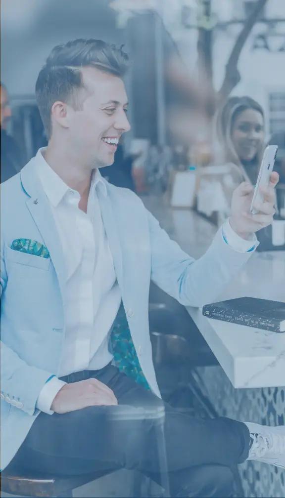 A man dressed in a nice suit seated at a marble table looking at his phone with a pretty girl looking at him in the background
